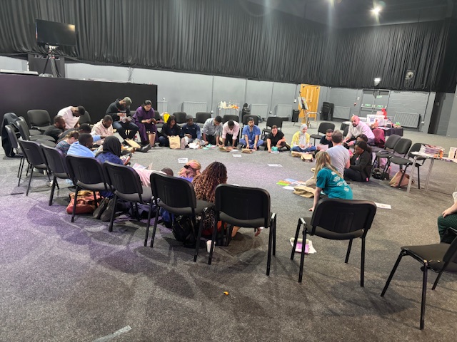 People at an event sat in a semi-circle, some on chairs and some on the floor