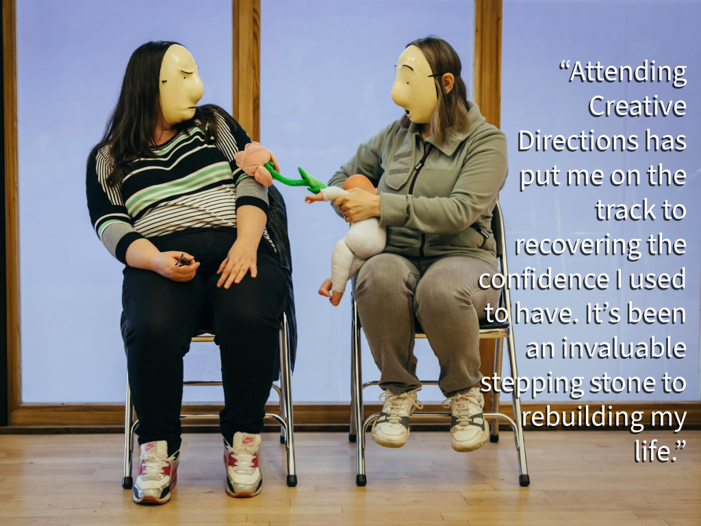 Two people sat on chairs. They have masks on their faces and are interacting with one another.
