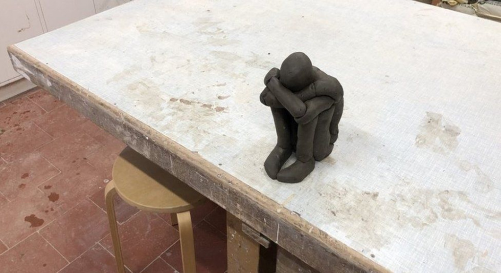 A model of a person sat crouched on the floor and holding legs. The creator is Sir Anthon Gormley and the piece is called 'Hold'.